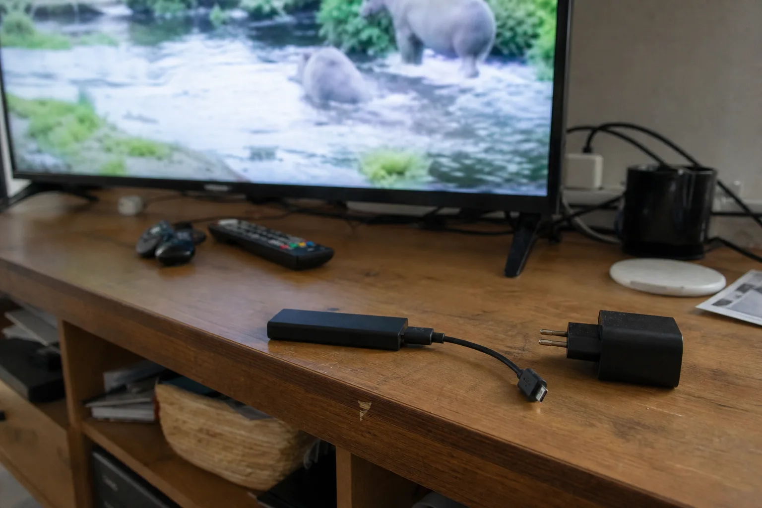 Fire TV‑Stick met losgekoppelde USB‑voedingskabel naast de adapter, tv op de achtergrond speelt een natuurdocumentaire.