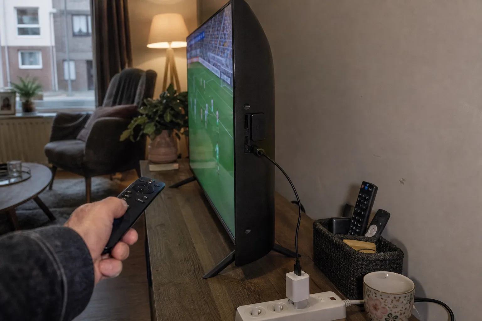 Streaming stick in TV HDMI with USB power to wall adapter; hand holding remote; football on screen.
